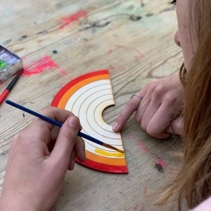 May include: A person paints a wooden rainbow with yellow paint using a blue paintbrush. The rainbow has red, orange, yellow, and white stripes.