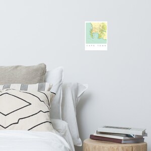May include: A rectangular print of a map of Cape Town, South Africa, in shades of green, yellow, and brown. The text "CAPE TOWN" is printed in white below the map on a light blue background. The print is displayed on a white wall.