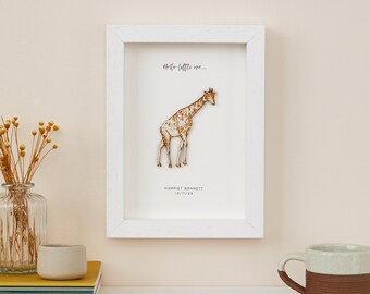 May include: A framed print featuring a giraffe illustration in shades of brown and cream. The white frame surrounds the artwork, which includes the text "Hello little one..." and a name and date. The print is displayed on a white shelf.