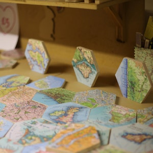 May include: A collection of hexagonal tiles featuring detailed map designs. The tiles display various geographical regions with a mix of colors, including blues, greens, and browns. Some tiles are standing upright, while others are laid flat, creating a textured surface.