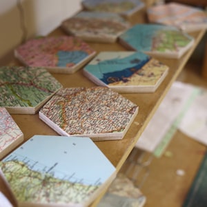 May include: A collection of hexagonal coasters with vintage map designs. The coasters feature various colors and map details, arranged on a wooden shelf. Each coaster has a unique map pattern, showcasing different regions.