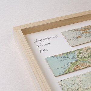 May include: A framed artwork featuring vintage map strips. The artwork includes the text "Happy Memories Were Made Here...". The frame is made of light-colored wood. The maps are in shades of blue, green, and tan.