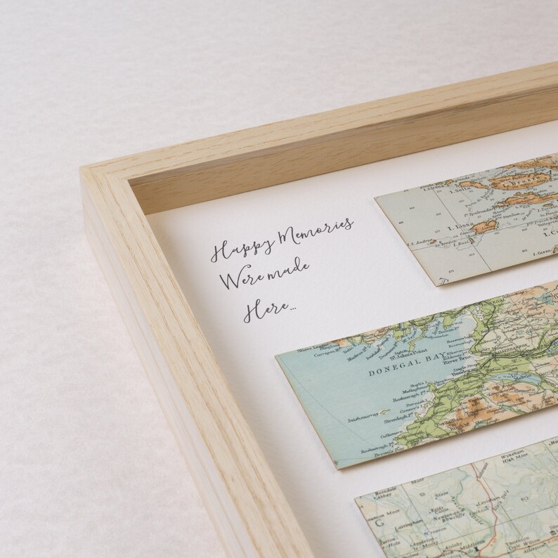 May include: A framed artwork featuring vintage map strips. The artwork includes the text "Happy Memories Were Made Here...". The frame is made of light-colored wood. The maps are in shades of blue, green, and tan.