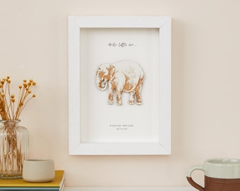 May include: White framed artwork featuring a wooden elephant cutout. The artwork includes the text "Hello little one..." and a name and date. The elephant is light brown and white, set against a white background.