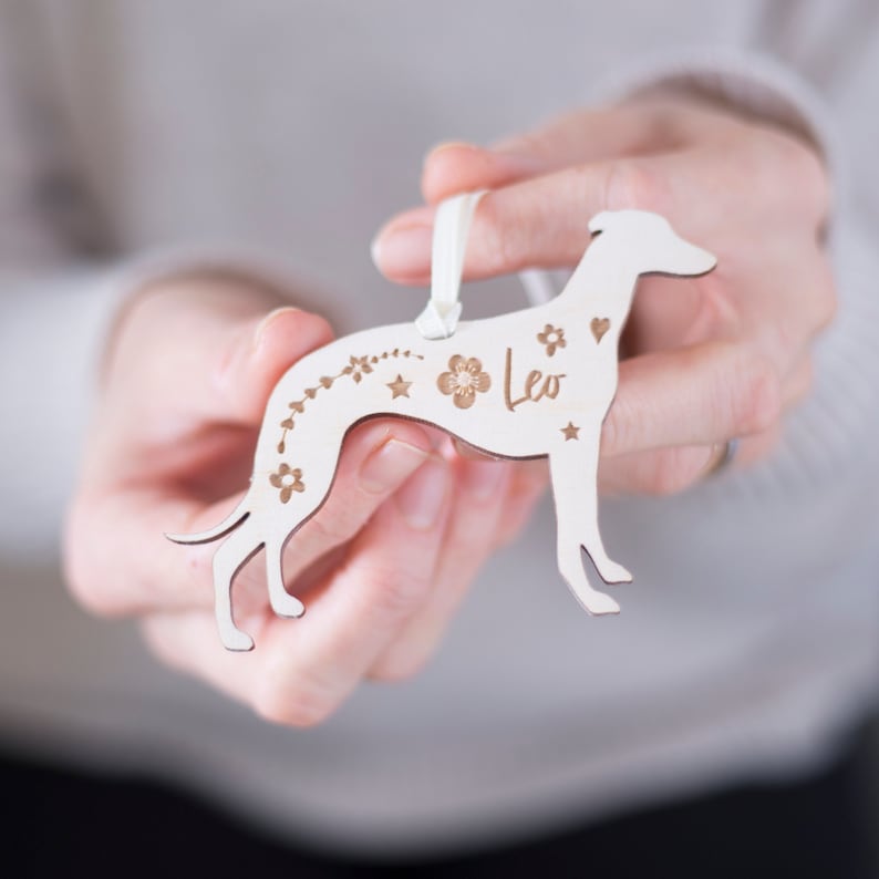 May include: White wooden ornament shaped like a Greyhound dog with the name "Leo" engraved on it. The ornament is decorated with small flowers and stars.