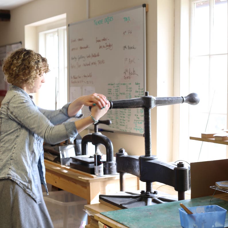 May include: A woman operates a large, antique printing press in a sunlit workshop. The press is made of dark metal and has a long lever. A whiteboard with handwritten notes is in the background. The scene suggests a focus on traditional craftsmanship.