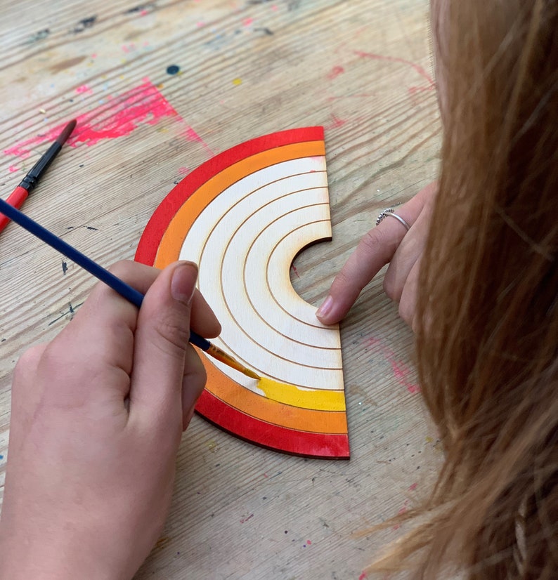 May include: A person paints a wooden rainbow with yellow paint using a blue paintbrush. The rainbow has red, orange, and yellow stripes.