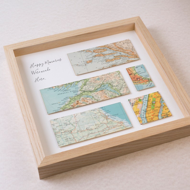 May include: A framed artwork featuring vintage map fragments. The maps, in shades of blue, green, and tan, are arranged on a white background within a light wood frame. The text "Happy Memories Were Made Here" is handwritten in black.