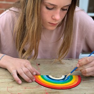 May include: A person paints a wooden rainbow with a blue paintbrush. The rainbow is made of multiple layers of wood, each painted a different color of the rainbow.