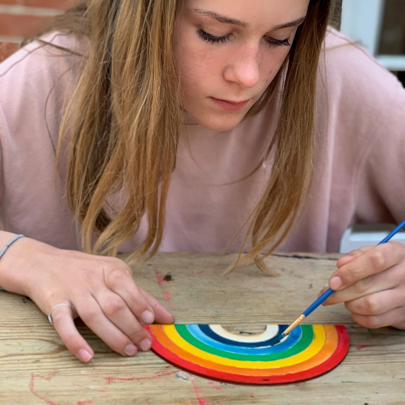 May include: A person paints a wooden rainbow with a blue paintbrush. The rainbow is made of multiple layers of wood, each painted a different color of the rainbow.