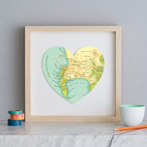 May include: A framed heart-shaped map of Cape Town, South Africa, with the city's name and surrounding areas labelled. The map is printed on a light blue background with green and brown accents. The frame is made of light wood.