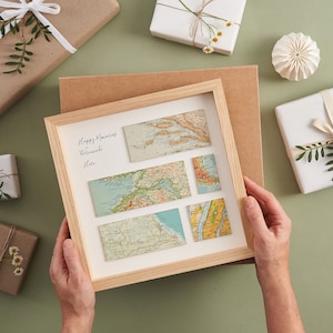 May include: A framed artwork featuring vintage map pieces arranged on a cream background. The wooden frame surrounds the maps, with handwritten text in the upper left corner. The maps are in shades of blue, green, and tan. Gift-wrapped presents surround the artwork.