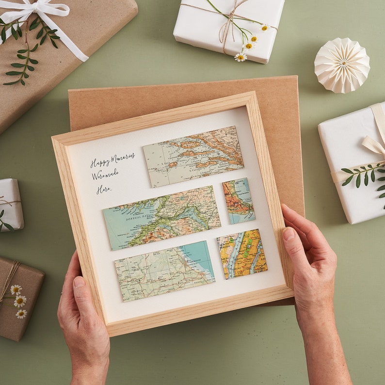 May include: A framed artwork featuring vintage map pieces arranged on a cream background. The wooden frame surrounds the maps, with handwritten text in the upper left corner. The maps are in shades of blue, green, and tan. Gift-wrapped presents surround the artwork.