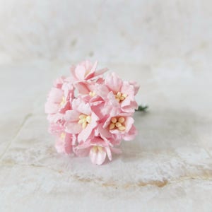 May include: A small bouquet of delicate pink paper flowers with yellow centers. The flowers are clustered together, creating a full, rounded shape. The stems are green. Ideal for crafting or adding a touch of floral charm.