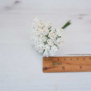 May include: A bunch of small white paper roses with green stems, measuring approximately 2 inches in diameter, on a white wooden surface.