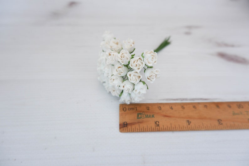 May include: A bunch of small white paper roses with green stems, measuring approximately 2 inches in diameter, on a white wooden surface.