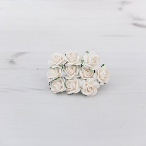 Puede incluir: Un grupo de pequeñas rosas de papel de color marfil con tallos verdes. Las rosas están dispuestas en un ramo suelto sobre una superficie de madera blanca.