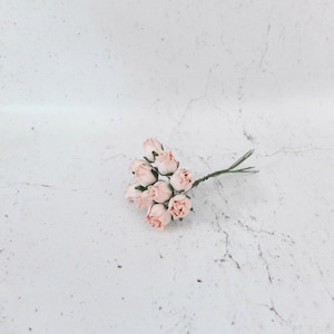 Pode incluir: Um ramo de pequenas rosas de papel rosa claro com caules verdes. As rosas estão dispostas em um cacho e são amarradas com um fio.