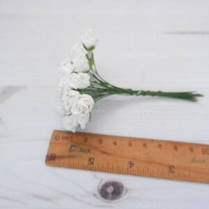 May include: A bunch of small white paper roses with green stems. The roses are arranged in a cluster and are about 2 inches in diameter.