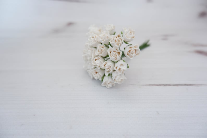 May include: A bunch of small, white paper roses with green stems. The roses are arranged in a cluster and are sitting on a white wooden surface.
