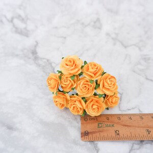 May include: A cluster of small, paper roses in a pale orange color. The roses are arranged in a bouquet and measure approximately 2 cm in diameter. The roses are on a white marble surface with a wooden ruler for scale.