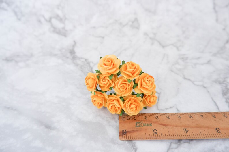 May include: A cluster of small, paper roses in a pale orange color. The roses are arranged in a bouquet and measure approximately 2 cm in diameter. The roses are on a white marble surface with a wooden ruler for scale.