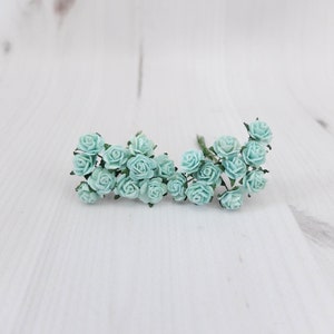 Puede incluir: Dos racimos de pequeñas rosas de papel azul claro. Las flores están dispuestas en un estilo suelto y natural, con tallos verdes. El fondo es una superficie blanca texturizada, creando una estética suave y delicada.