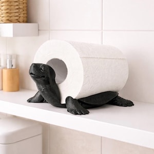 Op de afbeelding: Een zwarte toiletpapierhouder in de vorm van een schildpad. De kop en poten van de schildpad steken uit onder een rol wit toiletpapier. De houder staat op een witte plank in een badkamer.