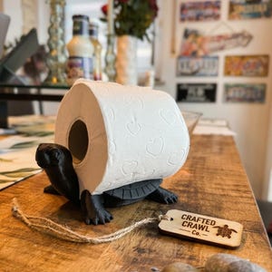 May include: A black turtle-shaped toilet roll holder supporting a roll of white toilet paper with a heart pattern. A wooden tag with the text "Crafted Crawl Co." is attached. The holder is on a wooden surface.