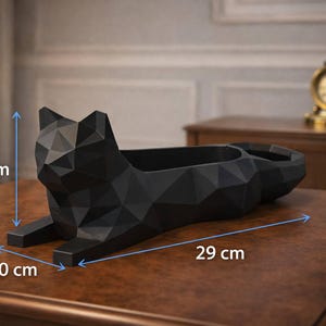 Könnte beinhalten: Ein schwarzer, geometrischer Katzen-Pflanztopf, 10 cm breit, 29 cm lang und 12 cm hoch. Der Pflanztopf hat ein Low-Poly-Design und besteht aus einem mattschwarzen Material. Er steht auf einer braunen Oberfläche.