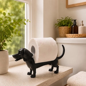 Könnte beinhalten: Ein schwarzer Toilettenpapierhalter aus Gusseisen in Form eines Dackels. Die Hundefigur steht auf vier Beinen mit nach oben gebogenem Schwanz und hat eine glatte, matte Oberfläche. Eine Rolle weißes Toilettenpapier liegt auf dem Rücken.