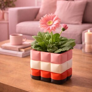 Könnte beinhalten: Eine rosa Gerbera in einem mehrfarbigen Pflanzgefäß. Der Pflanztopf ist mit rosa, weißen und orangefarbenen Blöcken gestapelt. Der Blumentopf steht auf einem Holztisch, im Hintergrund eine rosa Tasse und Bücher.