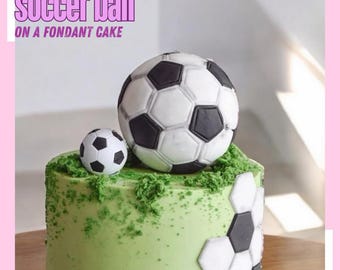 May include: A green fondant cake decorated with a soccer ball and smaller soccer ball on top. The cake is covered in green sprinkles and has a soccer ball pattern on the side.