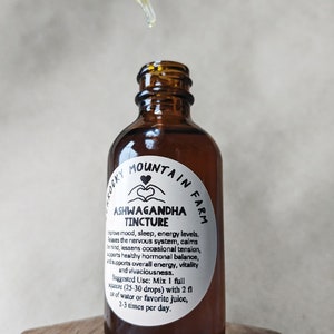 May include: A brown glass bottle with a white label that reads "Rocky Mountain Farm Ashwagandha Tincture". The label describes the benefits of the tincture, including improved mood, sleep, and energy levels. The bottle is sitting on a wooden surface.