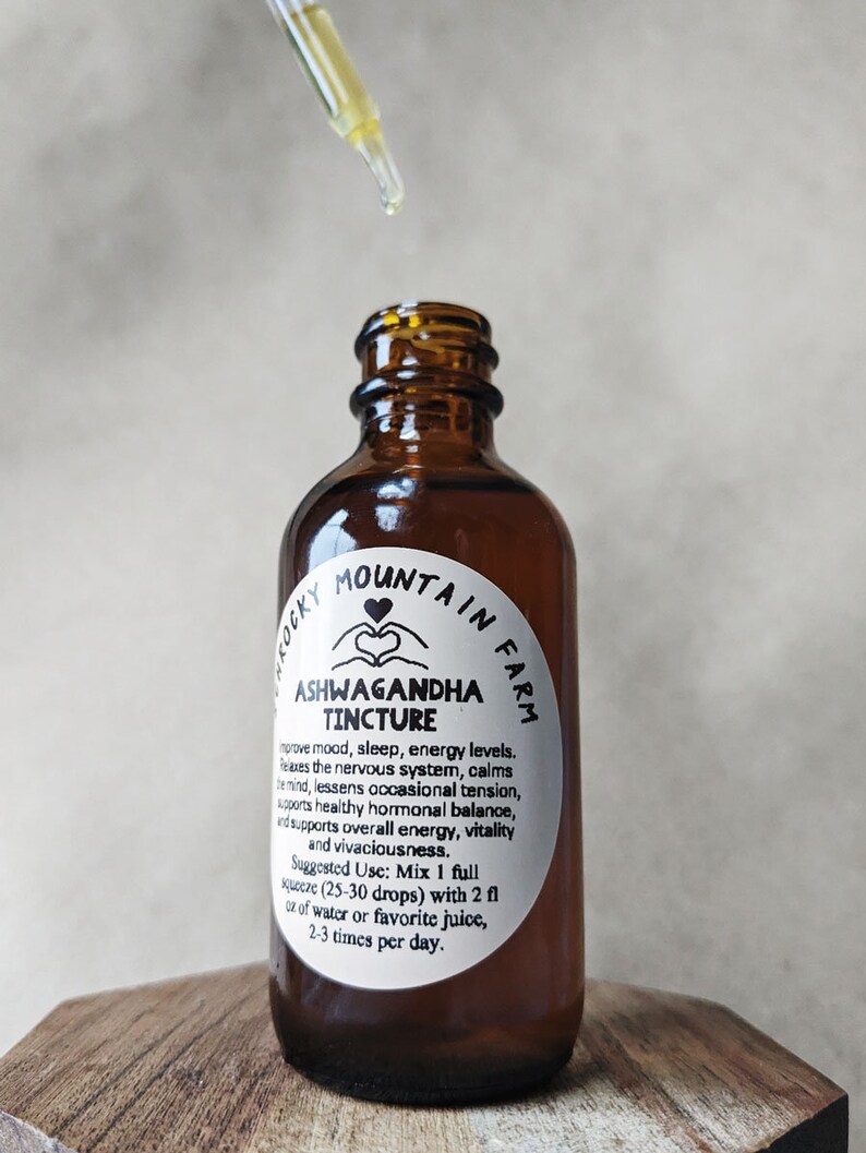 May include: A brown glass bottle with a white label that reads "Rocky Mountain Farm Ashwagandha Tincture". The label describes the benefits of the tincture, including improved mood, sleep, and energy levels. The bottle is sitting on a wooden surface.
