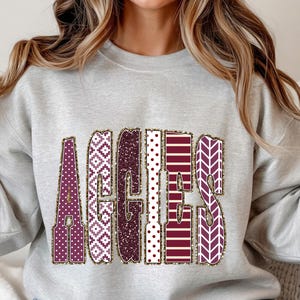 May include: A light gray sweatshirt with the word "AGGIES" in a decorative font. The letters are filled with various patterns in maroon, white, and gold, including polka dots, chevrons, and geometric designs. The sweatshirt has long sleeves.