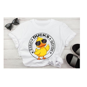 May include: White t-shirt with a graphic of a yellow duck wearing sunglasses, with the text "ZERO DUCKS GIVEN" in a circular design. Black sunglasses and sandals are visible.