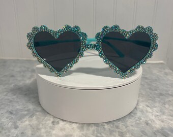 May include: Teal heart-shaped sunglasses with iridescent rhinestones. The sunglasses have dark lenses and a delicate, scalloped edge.