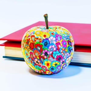 Könnte beinhalten: Eine dekorative Apfelskulptur, die mit bunten Blumenmustern bedeckt ist. Der Apfel hat einen braunen Stiel und steht vor roten Büchern. Die Blumen sind in Rot-, Gelb-, Blau- und Rosatönen gehalten.