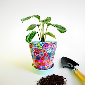May include: A vibrant, floral-patterned ceramic flower pot with a matching saucer, holding a green plant with large leaves. A small black trowel with a wooden handle and a pile of soil are next to the pot, suggesting gardening.