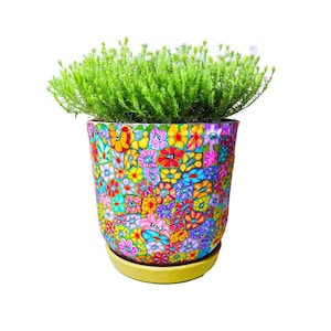 Könnte beinhalten: Ein farbenfroher Blumentopf mit einer leuchtend grünen Pflanze. Der Topf ist mit einem Mosaik aus Blumen in verschiedenen Farben verziert, darunter Rot, Gelb, Blau und Rosa. Der Topf steht auf einer passenden gelben Untertasse.
