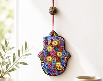May include: A hamsa hand wall hanging featuring a vibrant, multi-colored floral pattern in shades of red, yellow, blue, and purple. The ornament is suspended by a thin red cord adorned with a single matching floral bead at the top center.