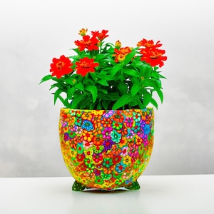 May include: A vibrant, colorful flower pot filled with bright red flowers and green foliage. The pot is adorned with a mosaic of small, multi-colored flowers, and rests on three small green feet. A cheerful, decorative item.