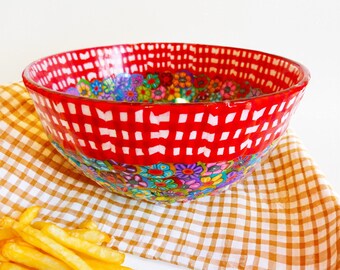 Pode incluir: Uma tigela de vidro decorativa com uma borda xadrez vermelha e branca e um padrão floral. A tigela é preenchida com flores coloridas e repousa sobre um pano xadrez. Uma pilha de batatas fritas está em primeiro plano.