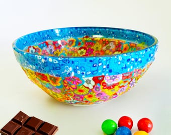 May include: A decorative bowl with a vibrant floral pattern in shades of yellow, pink, and blue. The rim is a solid blue with white dots. Chocolate squares and colorful gumballs are next to the bowl.
