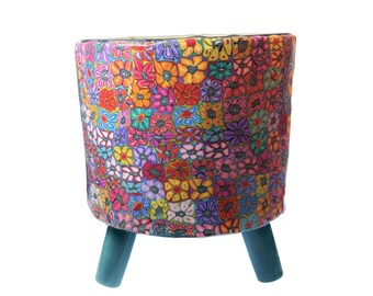 Könnte beinhalten: Ein zylindrischer Übertopf mit einem lebendigen, mehrfarbigen Blumen-Mosaikmuster in Rot-, Orange-, Gelb-, Lila- und Blautönen. Der Übertopf wird von drei massiven, glatten, blaugrünen Beinen getragen, die aus der Basis herausragen.