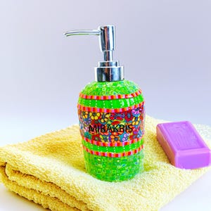 Puede incluir: Un dispensador de jabón de mosaico verde con una bomba plateada, adornado con coloridos diseños florales y el texto "MIRAKBIS". Una pastilla de jabón morado descansa sobre una toalla amarilla. El dispensador mide unos 15 cm de alto.