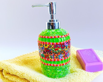 May include: A green mosaic soap dispenser with a silver pump, decorated with colourful floral patterns and the text "MIRAKBIS." A purple bar of soap sits on a yellow towel. The dispenser is approximately 15 cm tall.