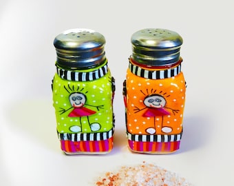 May include: Two decorative salt and pepper shakers with silver lids. One shaker is green with white polka dots, and the other is orange with white polka dots. Each has a painted cartoon figure and a striped base. A small pile of pink salt sits in front.