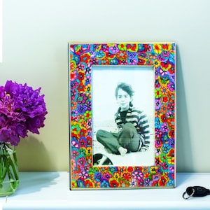 May include: A colorful picture frame with a floral design in shades of red, blue, yellow, and purple. The frame surrounds a black and white photograph of a person. A vase of purple flowers sits to the left.